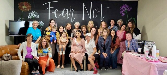 Un salón lleno de mujeres reunidas en un baby shower frente a una pared que dice Fear Not